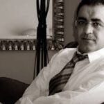 شهاب زريوح يجلس مبتسماً يرتدي نظارة وربطة عنق شبه رسمية