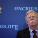 جون بولتون يبتسم أمام لافتة CRiUS