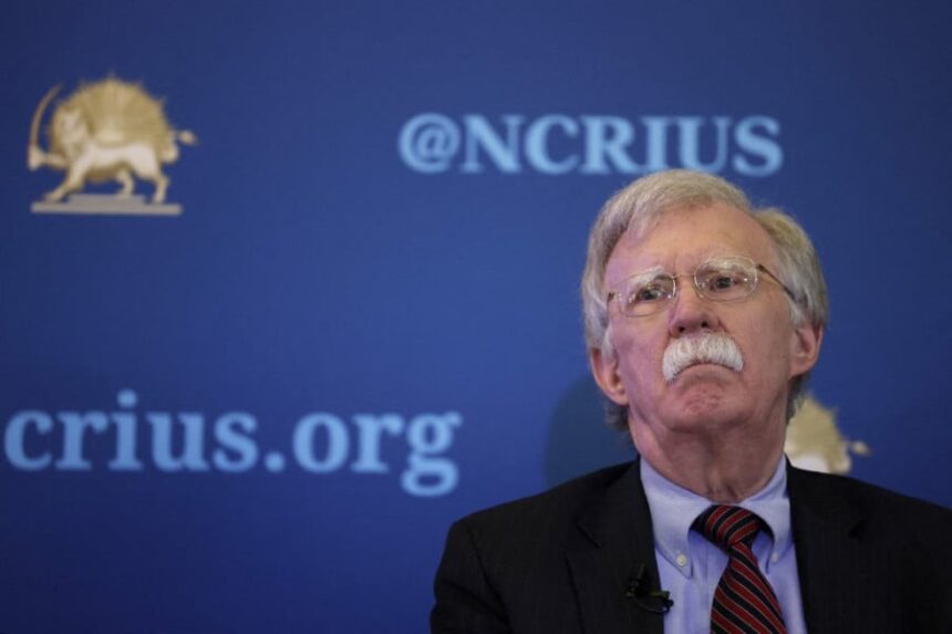 جون بولتون يبتسم أمام لافتة CRiUS