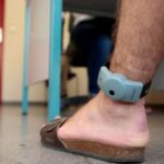 رجل بقدمه جهاز تتبع إلكتروني، وخلفه شخص يقف بجانب الطاولة.