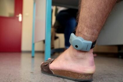 رجل بقدمه جهاز تتبع إلكتروني، وخلفه شخص يقف بجانب الطاولة.