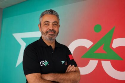 شاب بقميص أسود يحمل علامة CRA يقف مبتسماً أمام لافتة