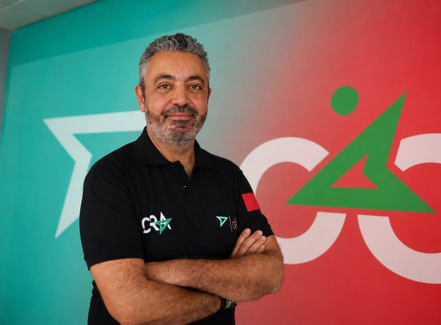 شاب بقميص أسود يحمل علامة CRA يقف مبتسماً أمام لافتة