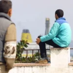 أشخاص يقفون ويجلسون على رصيف، سفن وسماء غائمة في الخلفية.