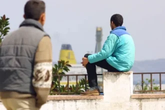 أشخاص يقفون ويجلسون على رصيف، سفن وسماء غائمة في الخلفية.