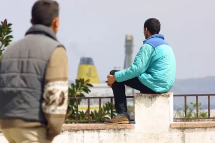 أشخاص يقفون ويجلسون على رصيف، سفن وسماء غائمة في الخلفية.