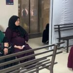 ثلاث نساء يجلسن على مقاعد خشبية رمادية داخل مستشفى في غزة.