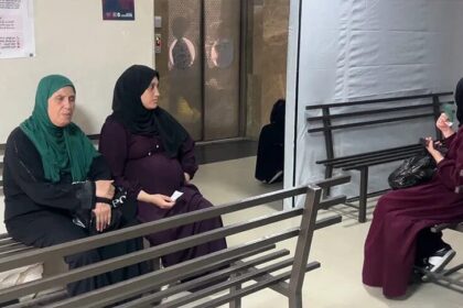 ثلاث نساء يجلسن على مقاعد خشبية رمادية داخل مستشفى في غزة.