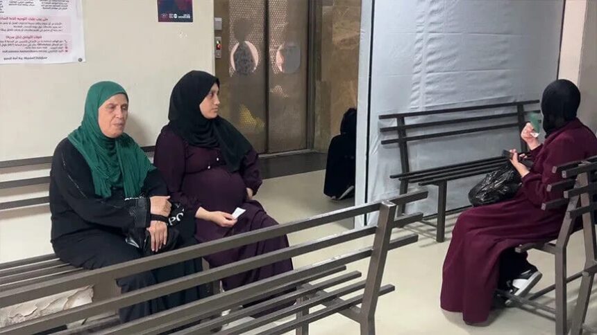 ثلاث نساء يجلسن على مقاعد خشبية رمادية داخل مستشفى في غزة.