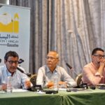 ثلاثة رجال يتحدثون أمام الميكروفونات في اجتماع.