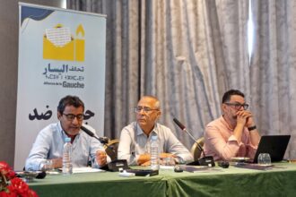 ثلاثة رجال يتحدثون أمام الميكروفونات في اجتماع.
