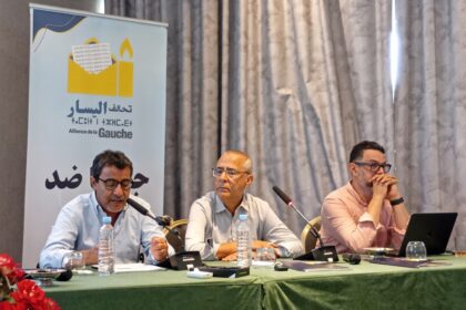 ثلاثة رجال يتحدثون أمام الميكروفونات في اجتماع.