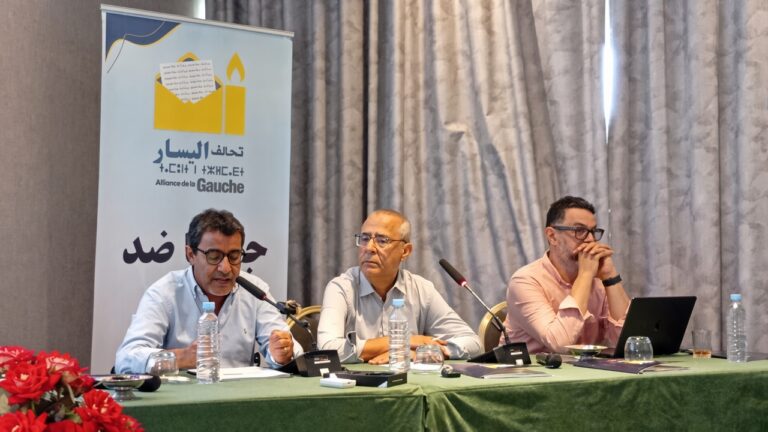 ثلاثة رجال يتحدثون أمام الميكروفونات في اجتماع.