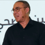 رجل يرتدي نظارة أمام الميكروفونات يتحدث